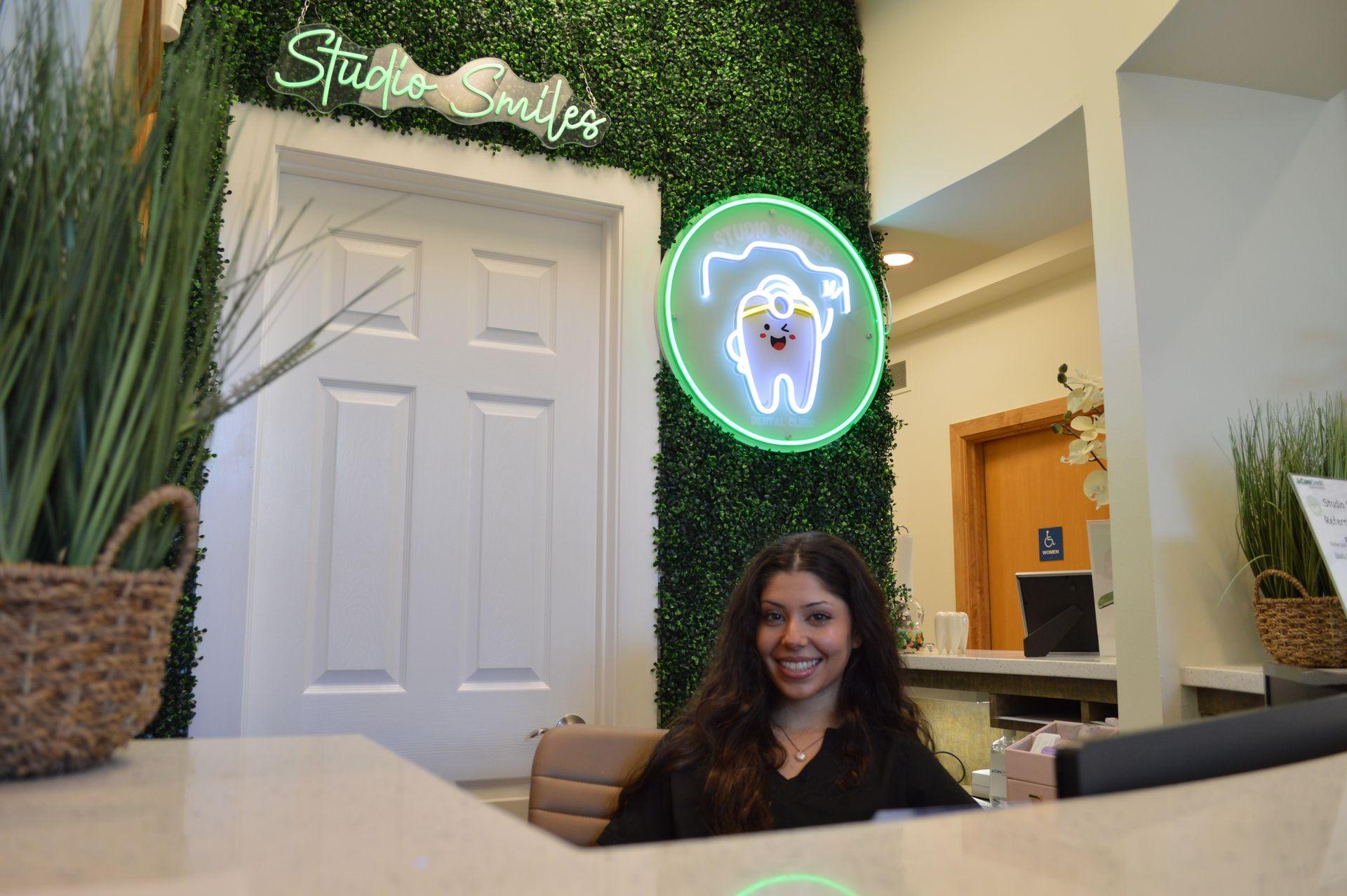 Reception area of a dental clinic with a smiling receptionist and neon tooth sign on a greenery wall.